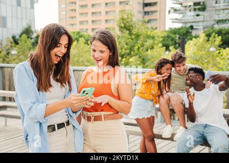 Two attractive and cheerful smiling women looking and showing the last funny and entertaining news on their smartphone outside at the university campus. At background, three young friends, having fun taking selfies in free time. Lifestyle concept. High quality photo Stock Photo