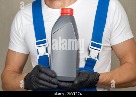 Close up auto mechanic in a white T-shirt and blue overalls holds a bottle of engine oil in front of him and looks into the camera, copy space. Stock Photo