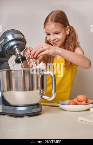 A positive girl of 9-10 years cooks homemade cake in the kitchen, beats eggs in a mixer on the kitchen table. Against the background of a white-gray Stock Photo