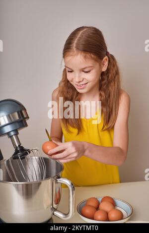 A positive girl of 9-10 years cooks homemade cake in the kitchen, beats eggs in a mixer on the kitchen table. Against the background of a white-gray Stock Photo