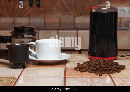 Preparing coffee: coffee beans, electric coffee grinder, coffee maker horn and white cup and saucer on a tiled kitchen table Stock Photo