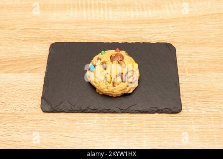 Delicious fluffy cookie with colored chocolate chips on a black slate plate on an oak table Stock Photo