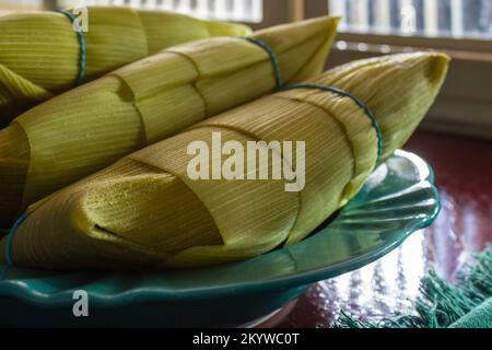 Goiânia, Goias, Brazil – December 01, 2022: 3 pamonhas covered with corn husks, inside a bright green plate next to a window. Stock Photo