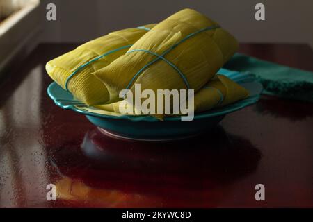 Goiânia, Goias, Brazil – December 01, 2022: 3 pamonhas covered with corn husks, inside a green dish, illuminated by the light from the window. Stock Photo