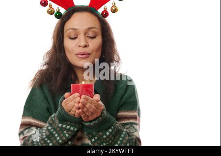 Attractive Latin American woman in warm green sweater and deer antler hoop, makes cherished wish while blows out candle Stock Photo