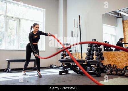 Battle ropes session. Attractive young slim and fit sportswoman working out in the gym for functional training doing crossfit exercises with battle ro Stock Photo