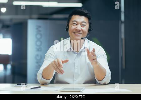 Online customer service. A young Asian man sits in the office in headphones and talks, consults remotely through a microphone, looks at the camera, smiles. Stock Photo