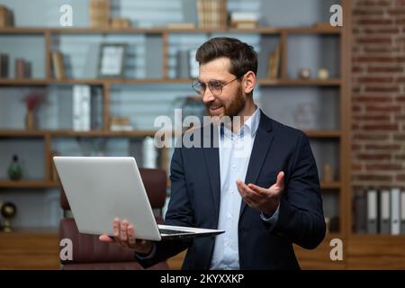 Video call of businessman, mature man standing in office and holding laptop talking remotely with colleagues, investor in business suit and glasses inside building. Stock Photo