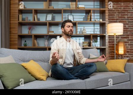 Mature man at home sitting on sofa in evening meditating in lotus position in living room. Stock Photo