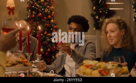 Happy multi cultural family celebrating Christmas or New Year, raising wineglasses. Served holiday table with different dishes and candles. Warm atmosphere of family Christmas dinner at home. Cheers. Stock Photo