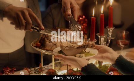 Close-up shot of African American man cutting turkey or chicken. Multi cultural family celebrating Christmas or Thanksgiving Day. Served table with dishes and candles. Family Christmas dinner at home. Stock Photo