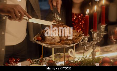 Close-up shot of African American man cutting turkey or chicken. Multi cultural family celebrating Christmas or Thanksgiving Day. Served table with dishes and candles. Family Christmas dinner at home. Stock Photo