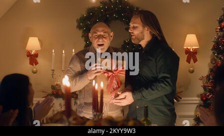 Man giving gift to his senior father. Happy family celebrating Christmas or New Year 2023. Served holiday table with candles. Warm atmosphere of family Christmas dinner at home. Stock Photo