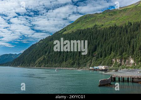 Seward, Alaska, USA - July 22, 2011: Green forested flank of mountain drops into azure Resurrection bay at south end of town under blue cloudscape. Qu Stock Photo