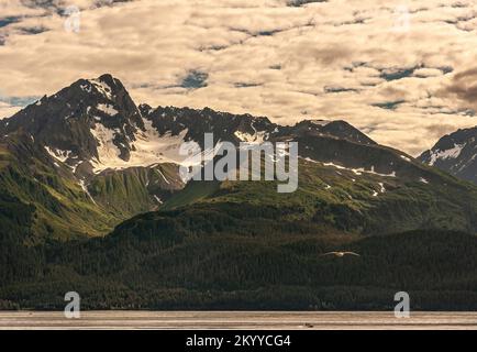 Seward, Alaska, USA - July 22, 2011: Closeup, under white cloudscape of dark mountain with snow patch in crevice and green forest on lower flanks. Fly Stock Photo