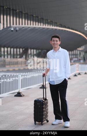 Asian young traveller with trolley case standing outside airport terminal, smiling at camera Stock Photo