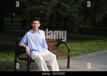 smiling handsome Asian young man, sitting on bench in park on sunny day. Looking at camera Stock Photo