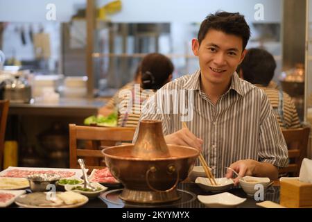 handsome Asian young man eating traditional Chinese hotpot in restaurant, looking at camera Stock Photo