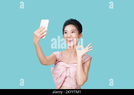 Happy young woman making silly faces while taking selfie on smartphone app with filters, standing against blue background Stock Photo