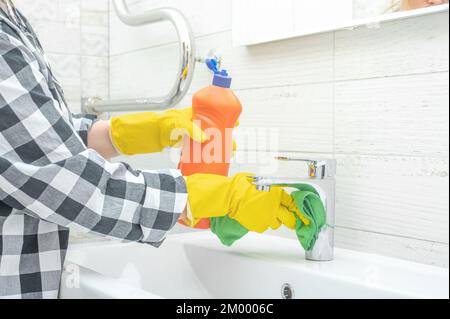 Cleaning Tap. Man doing chores cleaning bathroom at home. Cropped view of woman in rubber gloves with wet rag and detergent cleaning sink in bathroom. Stock Photo