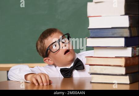 Who said school is easy. A young boy wearing glasses and a bow-tie looking wearily at a large pile of books. Stock Photo