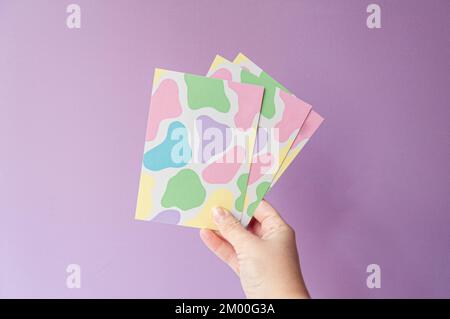 postcards with a funny cow design in pastel colors, held by female hand on lilac background. For ecommerce or small business Stock Photo