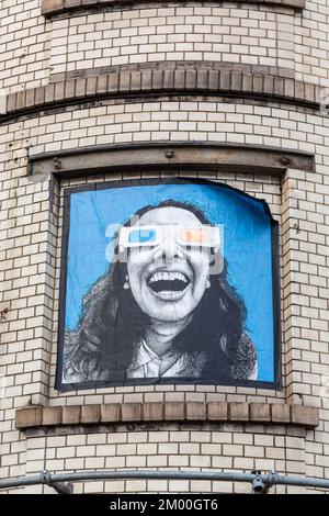 At a rounded corner of a light brick building, in a boarded-up window, image of a young woman wearing seroscopic vision goggles. Brussels. Stock Photo