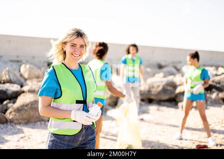 Young blonde woman smiling at the camera while her teamwork cleaning the rubish on a beach, volunteers collecting the waste on the coast line, young p Stock Photo