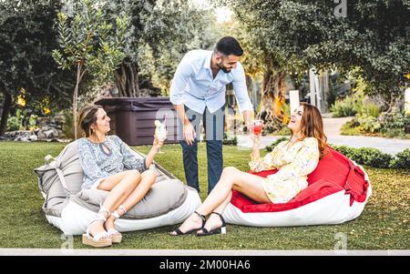 Two young women are served drinks by a male friend at a cocktail party in the garden, summer time lifestyle concept Stock Photo