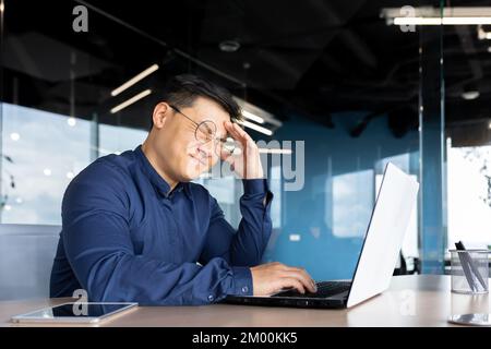 Asian businessman boss sick has severe headache, man in glasses sitting at desk using laptop at work inside office, migraine office worker investor overtired. Stock Photo