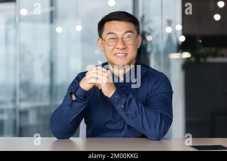 Young successful Asian businessman looking at caper and smiling, video call sitting at table investor boss in shirt and glasses. Stock Photo