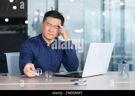 Asian businessman boss sick has severe headache, man in glasses sitting at desk using laptop at work inside office, migraine office worker investor overtired. Stock Photo