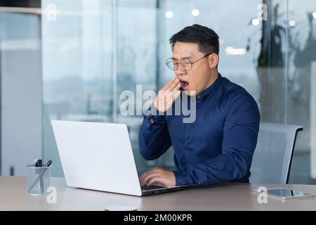 Tired worker at work yawning, asian man in shirt and glasses working inside office using laptop, man sleepless at work. Stock Photo