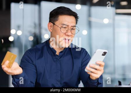 Cheated online buyer, asian businessman in office close up trying to make bank transaction and purchase in online store, man unhappy holding bank credit card and phone. Stock Photo