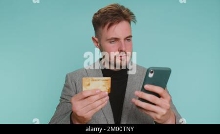 Sincere business man customer using credit bank card and smartphone while transferring money, purchases online shopping, payment. Finance and internet. Young guy isolated alone on blue wall background Stock Photo