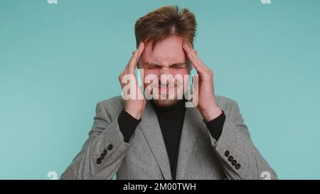 Bearded young adult business man rubbing temples to cure headache problem, suffering from tension and migaine, stress, grimacing in pain, high blood pressure isolated alone on blue wall background Stock Photo