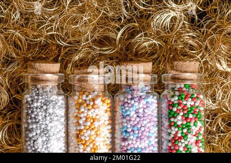 Nonpareils in small glass bottles, over golden angel hair. Winter and christmas colored mixes of edible confectionery of tiny sugar balls. Stock Photo