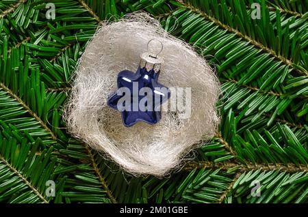 Blue glass star, in a silvery-white angel hair nest, over fresh green fir branches. Star shaped glass bauble to hang on a Christmas tree. Stock Photo