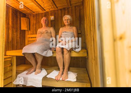 Leisure activity and relaxation concept. Full-length indoor shot of two white grandparents who sit in a wooden sauna with towels under their feet. High quality photo Stock Photo