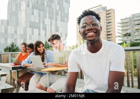 On foreground a smiling cheerful young African american student with a positive smile, white perfect teeth, looking at camera sitting outside taking a break at the university. at the background a group of university students have meet to research and share information about their homeworks using laptops and smatphones together in a positive environment. Education concept. High quality photo Stock Photo