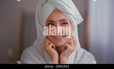 Close up portrait old middle-aged mature senior Caucasian smiling woman face lady in bath robe with towel on head with skincare eyes patches for Stock Photo