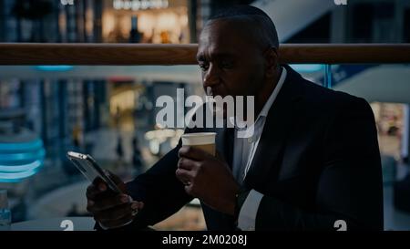 Middle-aged African entrepreneur ethnic man worker businessman sitting in cafe cafeteria looking at mobile phone using Wi-Fi browsing working with Stock Photo
