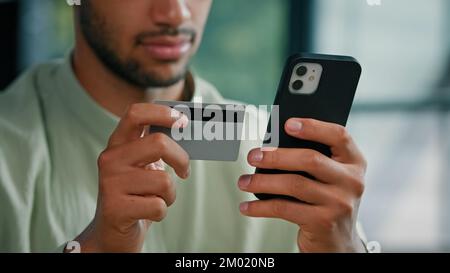 African American bearded man indoors unrecognizable male customer holding credit card and smartphone sitting at home office using easy mobile payment Stock Photo