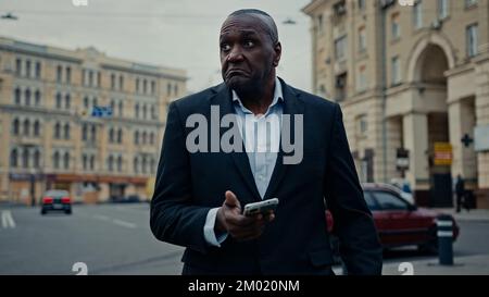 Ethnic adult man African middle-aged businessman entrepreneur with phone in city booking taxi with mobile app waiting car confused upset looking Stock Photo
