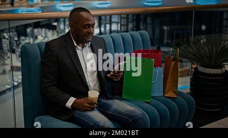 African adult man middle-aged businessman with shopping bags in mall store shop with coffee tea cup browsing scrolling phone mobile app buying online Stock Photo