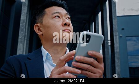 Close up mature 40s Korean businessman in city using mobile phone typing browsing booking taxi business chatting texting with smartphone Asian man Stock Photo
