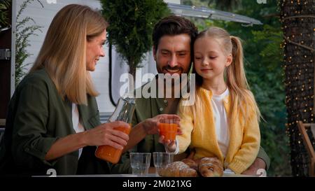 Caucasian family on camping vacation outdoors sitting near van have lunch picnic eat good with croissants mother pouring fresh orange juice into Stock Photo