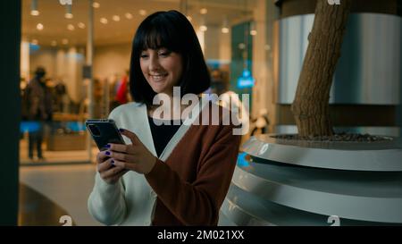 Smiling 20s woman lady female girl indoors in shopping mall sitting looking at mobile phone screen scrolling browsing in net app with smartphone smile Stock Photo