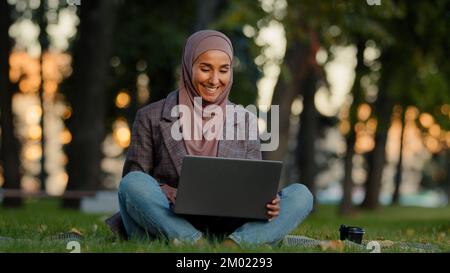Happy smiling muslim islamic business woman girl female student wears hijab sitting on green grass lawn in city park using laptop for distant e Stock Photo