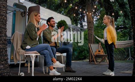 Caucasian mother and adult bearded father in camping near van trailer listening little daughter child girl kid singing song tell poem parents clapping Stock Photo